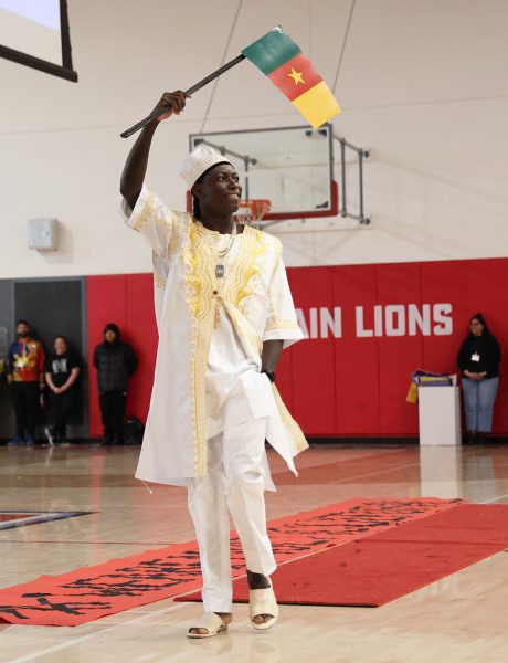 Senior Ynski Noel wears a traditional white and gold karabela to represent Haiti while he waves the flag of Cameroon high in the air. He smiles at the crowd on his left as he walks down the red carpet on the gym floor. Students dress in various outfits to represent their cultures during Multicultural Week. 
