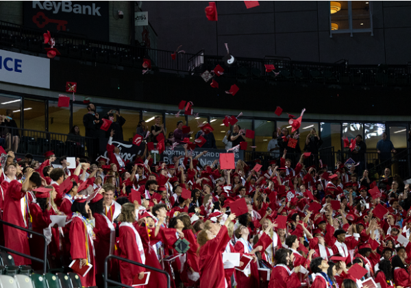 Students throw their caps into the air, after turning their tassels as a send off to their senior year. People can be seen in the back clapping and cheering as their caps fly.