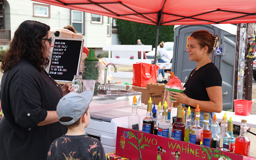 Wiebke can be seen handing a strawberry flavored shaved ice cone to her customers while smiling widely. A more detailed list of the shop's many flavors is posted to the left of the customer. Wiebke vocalized her fondness of the fair and how she appreciates all of the diversity it brings. 