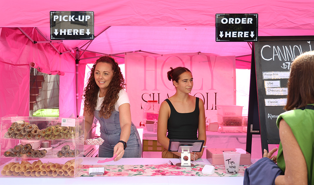 Shannon Smith (left), owner of Holy Shannoli, is assisted by Lainie Goldsand (right) behind the bright pink counter that marks the front of their booth. They sell a plethora of flavored cannolis, including pistachio, ube, cheesecake, espresso, strawberry, classic and many other seasonal flavorings.