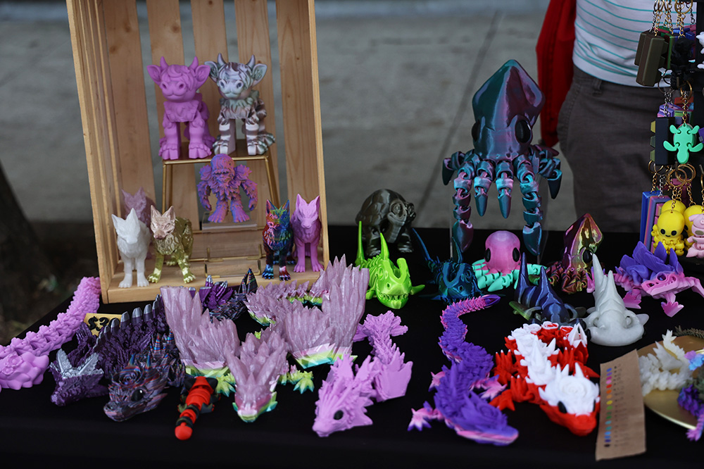 A multitude of 3D printed purple dragons crowd the left side of the table, while a red and white rose dragon lays to the right, next to multiple shark prints. Multi-colored cows and wolves are stacked in a box towards the back right portion of the table, while squids, octopi, turtles and fidgets adorn the back right side. A majority of the prints come in a variety of colors that the customers can choose from. 