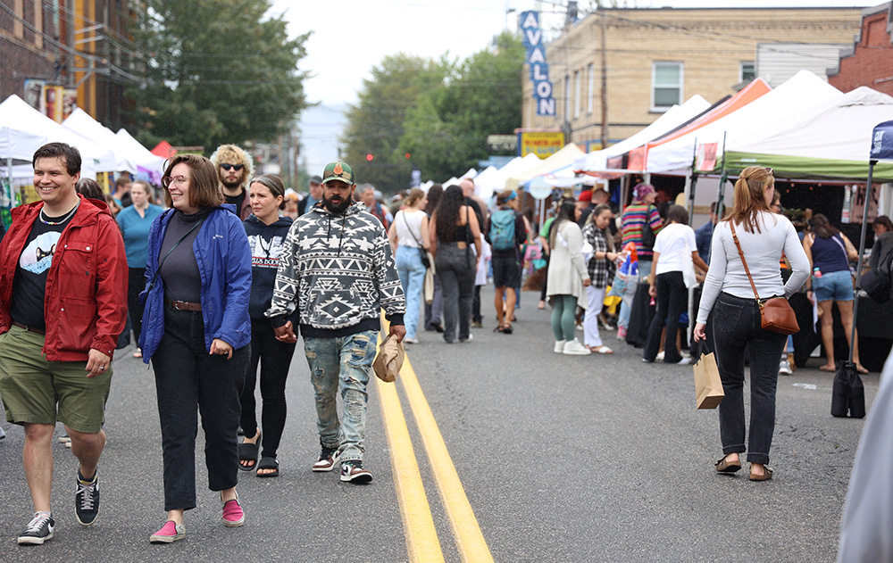 A crowd of people walk along Belmont Street, as they visit the vast amount of shops and booths open for the fair. Many of them smile widely as they walk with their friends and family, while others stop at shops along the street. The Belmont Street Fair brings together a diverse group of participants.