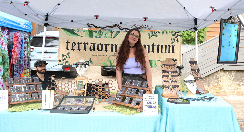 Terraornamentum, a jewelry company owned by Dinah Hunsicker, displays items such as micro terrarium jewelry, chokers, necklaces, earrings and pressed lichen. Hunsicker can be seen  standing behind the table exhibiting their merchandise, smiling towards the camera in a white tube top shirt and black choker. More information about this business can be found at terraornamentum.com. Hunsicker stated that they enjoy the good vibe and “laid back” feeling of the fair. 