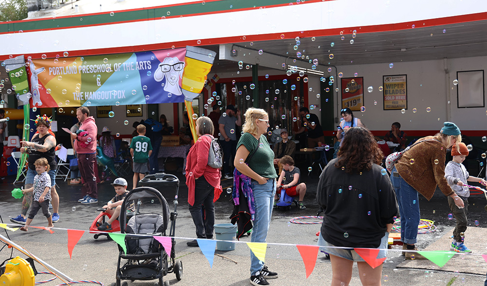 Many children can be seen taking part in activities such as blowing bubbles, riding kids scooters and even just walking or interacting with their parents and caretakers. Multi-colored bubbles float through the air, originating from the bubble machine to the left of the play area. The Portland Preschool of The Arts & Hangout PDX provides a place where even the youngest people can build community. 