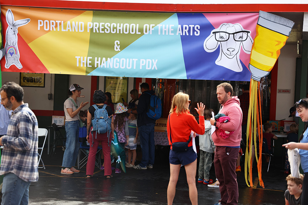 Portland Preschool of The Arts & Hangout PDX is a local place where kids can spend their time during the annual fair. Multiple adults and parents can be seen conversing with each other in front of the large, multicolored banner that includes a picture of a goat with black glasses. The kids zone is located in the parking lot of Battery X-Change on SE 38th and Belmont. 